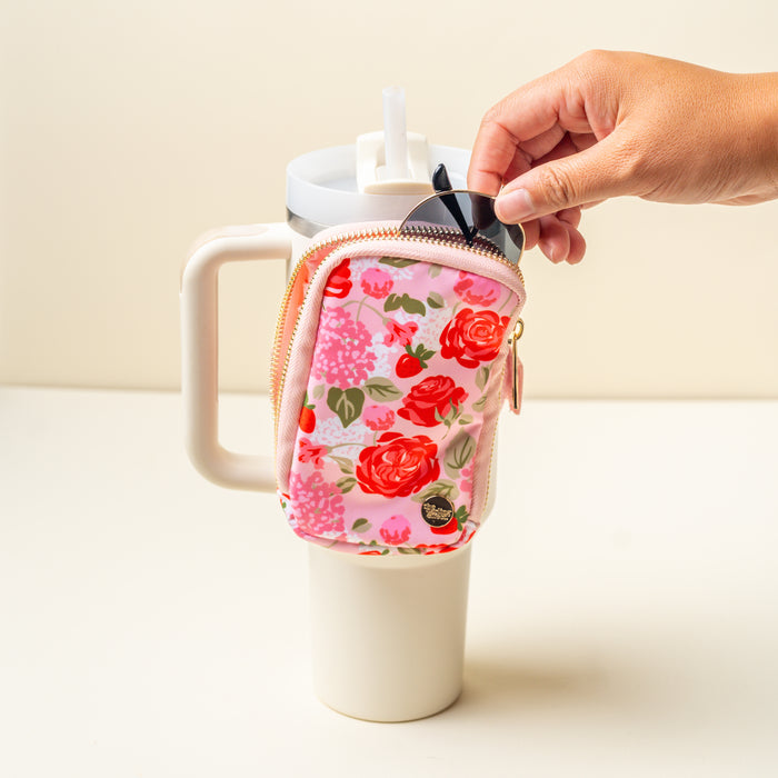 A hand unzips The Darling Effect Rose Garden Tumbler Fanny Pack attached to a large white tumbler with a straw, revealing a key fob partially tucked inside the stylish pink floral pouch.