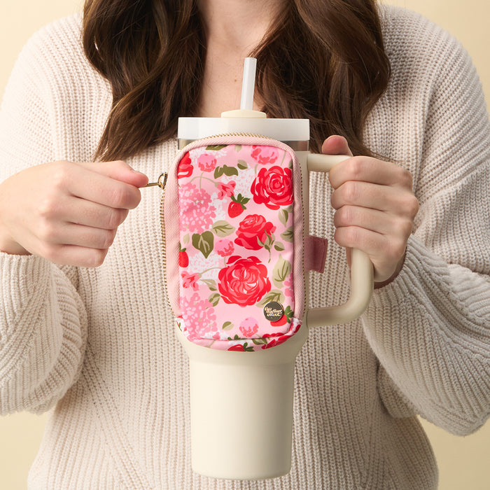 A woman in a cream sweater holds a travel mug with The Darling Effect's Rose Garden Tumbler Fanny Pack—a pink floral, water-resistant accessory—attached to the handle.