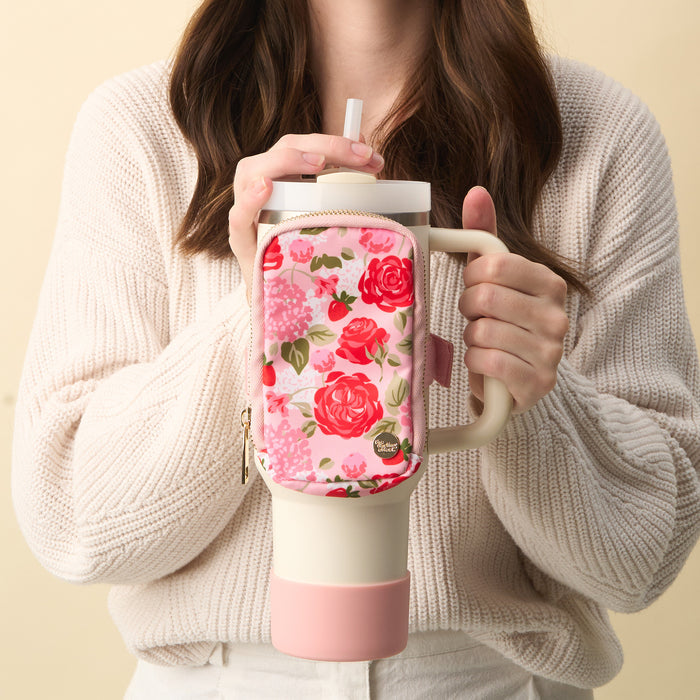 A woman in a cream sweater holds a large cup with a handle and straw, featuring The Darling Effect's Rose Garden Tumbler Fanny Pack—a water-resistant pink and floral zippered pouch attached to the front.
