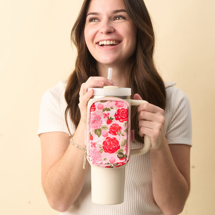 A smiling woman with long brown hair holds a large cream tumbler with a straw, featuring The Darling Effect's Rose Garden Tumbler Fanny Pack—a stylish pink floral accessory—attached to the handle. She wears a short-sleeved, ribbed white top.