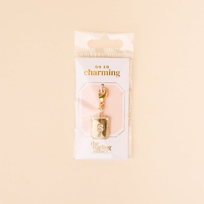 The-darling-effect S Jewelry Charm Initial Letter Gold, a gold-plated brass keychain with a rectangular pendant, is displayed in clear Oh So Charming packaging—an elegant, gift-ready piece set against a soft beige background.