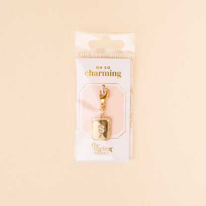 The-darling-effect S Jewelry Charm Initial Letter Gold, a gold-plated brass keychain with a rectangular pendant, is displayed in clear Oh So Charming packaging—an elegant, gift-ready piece set against a soft beige background.