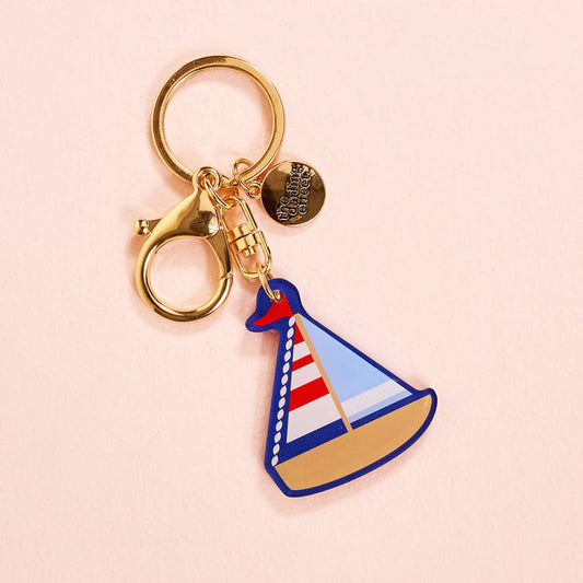 The Darling Effect’s Sailboat Acrylic Keychain features a gold keychain, blue sailboat charm with tan hull and red and white striped sail, plus a small gold disc with “the darling check” for extra charm.