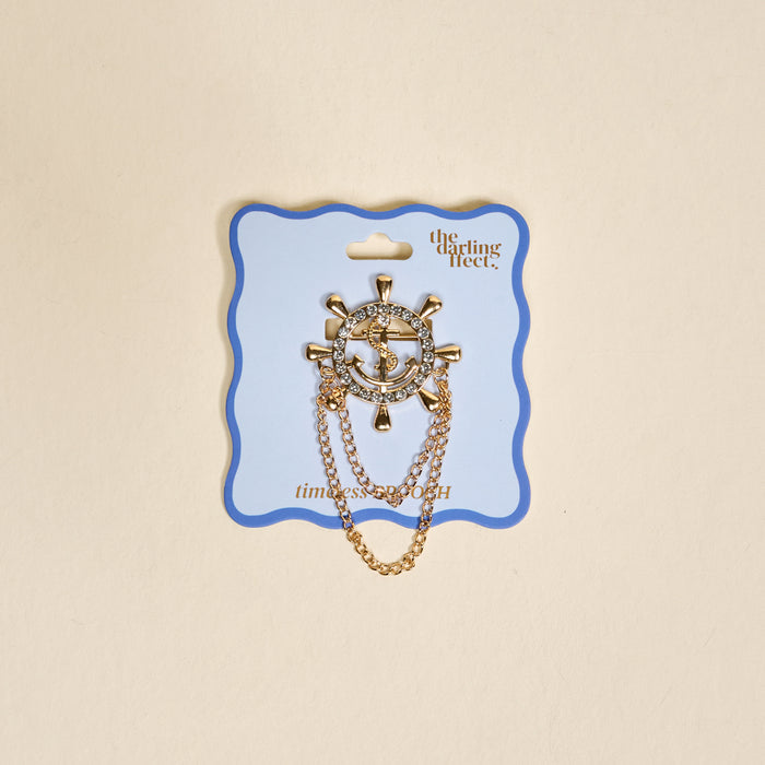 The Darling Effect’s Sailor's Charm Timeless Brooch features a vintage gold nautical design with a ship wheel, anchor, rhinestone accents, and two dangling chains, displayed on a scalloped blue and white card.