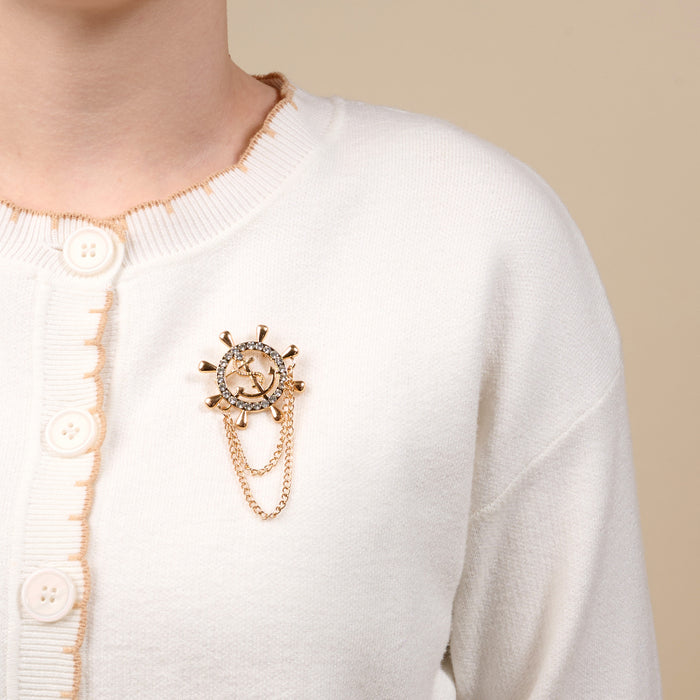The Darling Effect’s Sailor's Charm Timeless Brooch—a vintage-inspired gold ship’s wheel and anchor with diamond accents and dangling chains—adds elegance to any white cardigan and makes a memorable keepsake accessory.