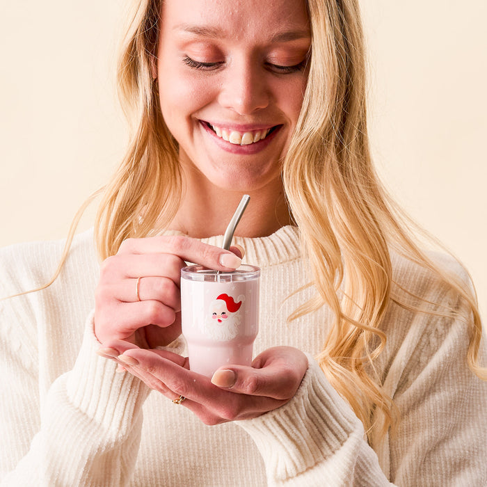 A smiling woman with long blonde hair holds The Darling Effect Santa 2 oz Tiny Tumbler, featuring a cute Santa face and filled with a pink drink and metal straw. She wears a cream sweater, looking down at the tumbler against a neutral background.