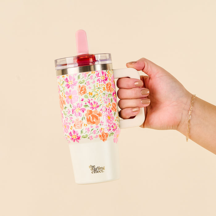 A hand holds the-darling-effect Secret Garden Pink 20 oz Lifestyle Flip Straw Tumbler, featuring a pink and orange floral design, pink straw, beige handle, and visible logo.