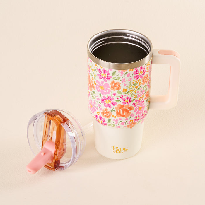 The Secret Garden Pink 20 oz Lifestyle Flip Straw Tumbler by the-darling-effect, featuring a floral pattern and pink handle, sits partially filled with a dark liquid. Its pink and transparent leak-resistant lid is placed nearby on the light surface.