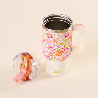 The Secret Garden Pink 20 oz Lifestyle Flip Straw Tumbler by the-darling-effect, featuring a floral pattern and pink handle, sits partially filled with a dark liquid. Its pink and transparent leak-resistant lid is placed nearby on the light surface.