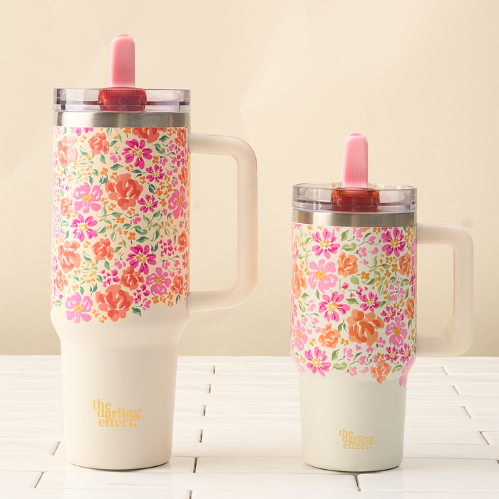 Two Secret Garden Pink 20 oz Lifestyle Flip Straw Tumblers by the-darling-effect, each floral-patterned with handles and pink straws, sit on white tiles. Both leak-resistant tumblers are printed with “the darling effect” in gold.