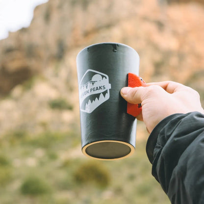 Hand holding Seven Peaks Climber 7a Mug near rocky hills, perfect outdoor coffee gift for mountain climbers and hikers.