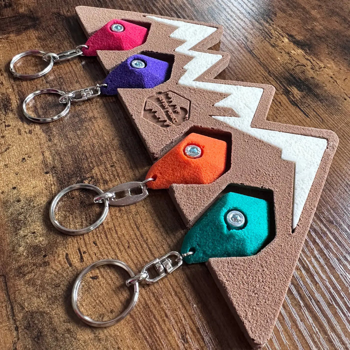 Three Seven Peaks climbing keychains in brown Rock Climber Holder on wood surface, perfect for outdoor adventure lovers.