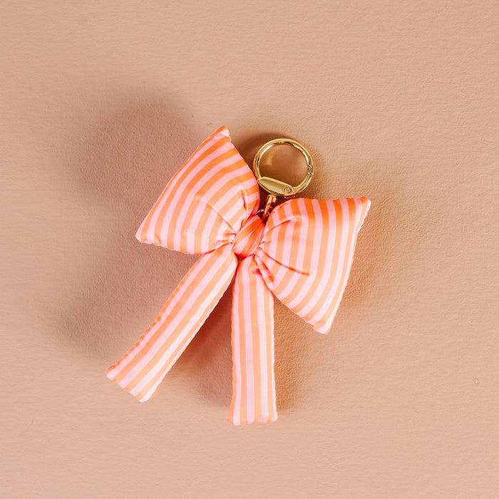 The Darling Effect's Sherbet Stripes Puffy Bow Bag Charm features a gold keychain and a soft pink and white striped bow, displayed on a light peach background—a perfect coquette accessory.