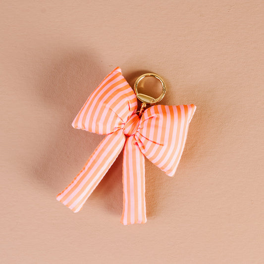 The Darling Effect's Sherbet Stripes Puffy Bow Bag Charm features a gold keychain and a soft pink and white striped bow, displayed on a light peach background—a perfect coquette accessory.