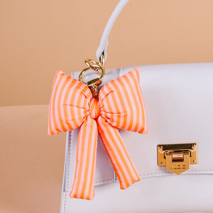 A close-up of a white handbag with a gold clasp and keychain featuring The Darling Effect’s Sherbet Stripes Puffy Bow Bag Charm—a large orange and pink striped bow with gold hardware—hanging playfully from the handle.
