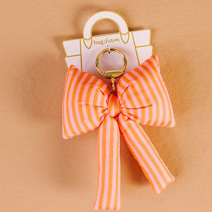 The Darling Effect’s Sherbet Stripes Puffy Bow Bag Charm, featuring pink and orange stripes with a gold keyring, is shown against a tan background—an ideal coquette accessory for your favorite purse.