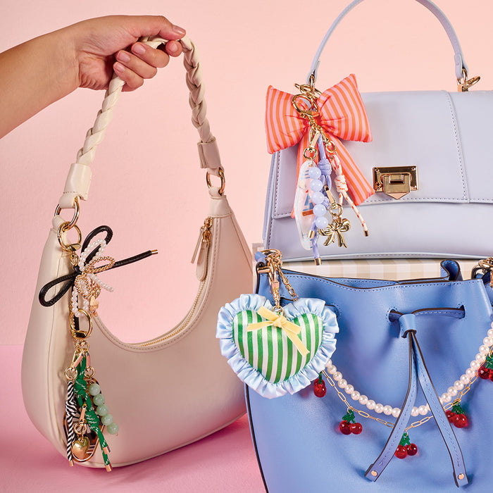 A hand holds a cream-colored purse with a braided strap. Behind it, light purple, light blue, and blue purses are stacked, each featuring The Darling Effect's Sherbet Stripes Puffy Bow Bag Charm and other coquette accessories.