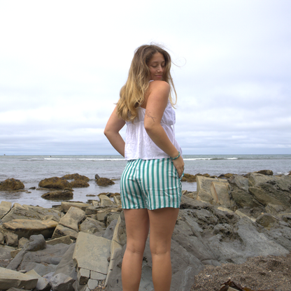 A woman with long hair stands on rocky terrain by the ocean, wearing a white sleeveless top and The Darling Effect's Sicily Stripes Teal All Day Boxer Shorts, made from breathable fabric, as she looks over her shoulder on a cloudy day.