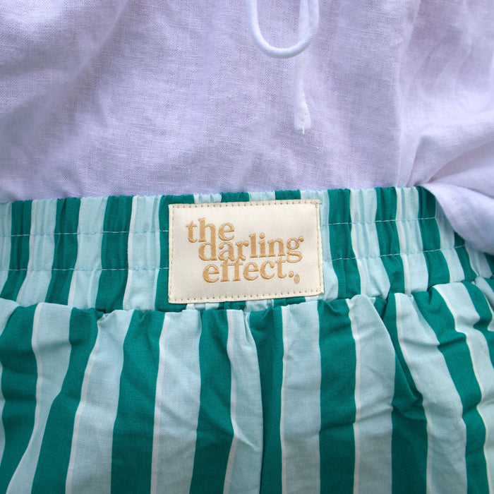 Close-up of Sicily Stripes Teal All Day Boxer Shorts by The Darling Effect, featuring green and white vertical stripes, breathable fabric, and a beige waistband label. A white garment is partially visible above the waistband.