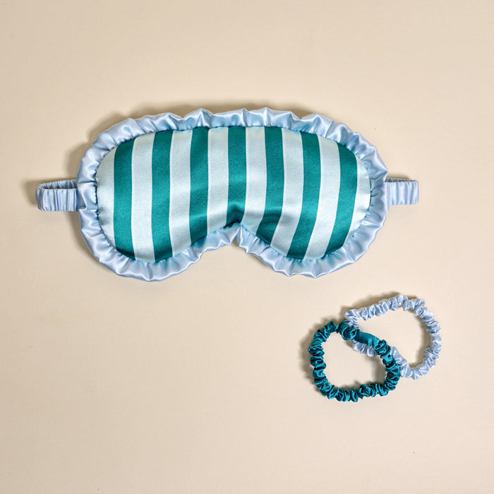 The Darling Effect’s Sicily Stripes Teal Beauty Sleep Mask, featuring soft satin fabric with light blue and teal vertical stripes and ruffled edges, comes with two matching scrunchies, shown on a beige background.