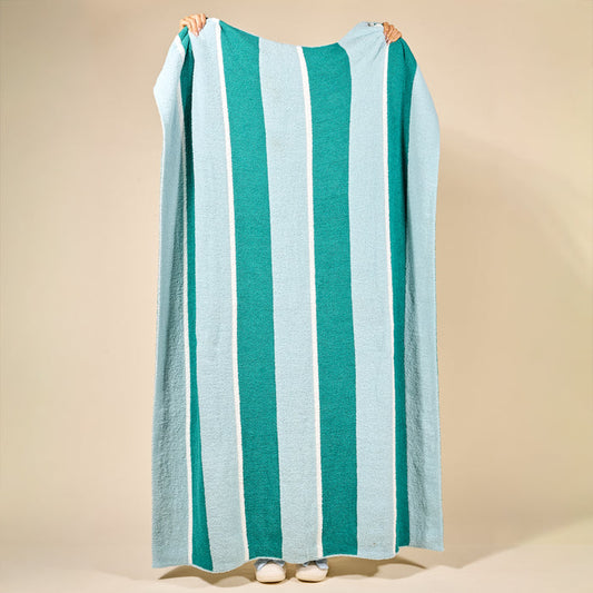 A person holds a large Sicily Stripes Teal Cozy Dreams Luxe Blanket by The Darling Effect, featuring vertical turquoise, light blue, and white stripes and crafted from ultra-soft microfiber for maximum comfort.