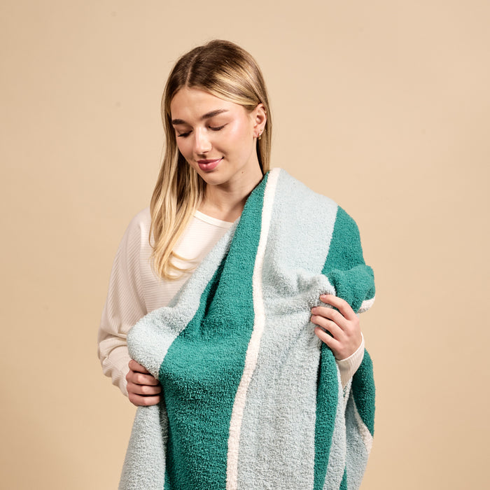 A young woman in a white sweater smiles while holding The Darling Effect Sicily Stripes Teal Cozy Dreams Luxe Blanket, featuring ultra-soft striped teal and light blue, against a beige background.