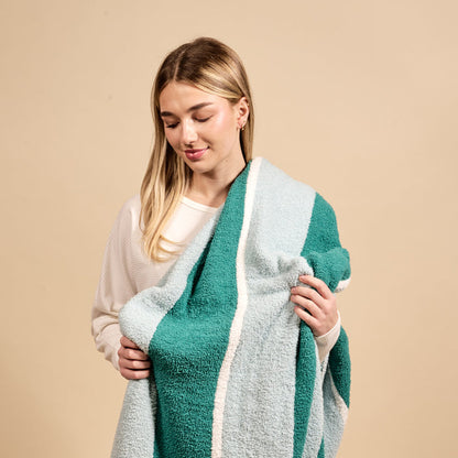 A young woman in a white sweater smiles while holding The Darling Effect Sicily Stripes Teal Cozy Dreams Luxe Blanket, featuring ultra-soft striped teal and light blue, against a beige background.