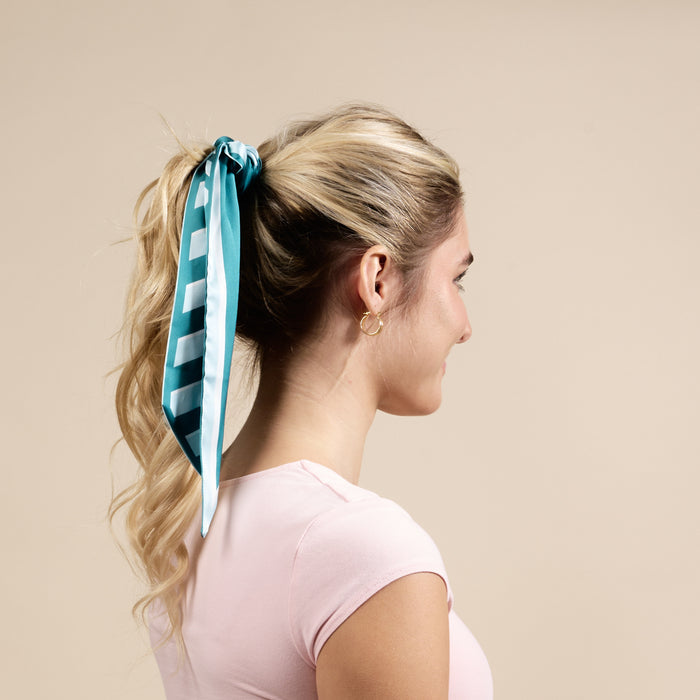 A woman with blonde hair in a ponytail, tied with The Darling Effect's Sicily Stripes Teal Twirl & Tie Scarf Accessory, wears a light pink shirt and gold hoop earrings, facing sideways against a plain beige background.