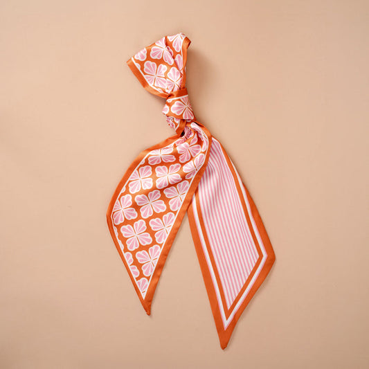 The Sienna Spice Twirl & Tie Scarf Accessory by The Darling Effect showcases vibrant orange and white floral and striped patterns, tied in a loose knot and displayed on a plain beige background, offering endless styling possibilities.