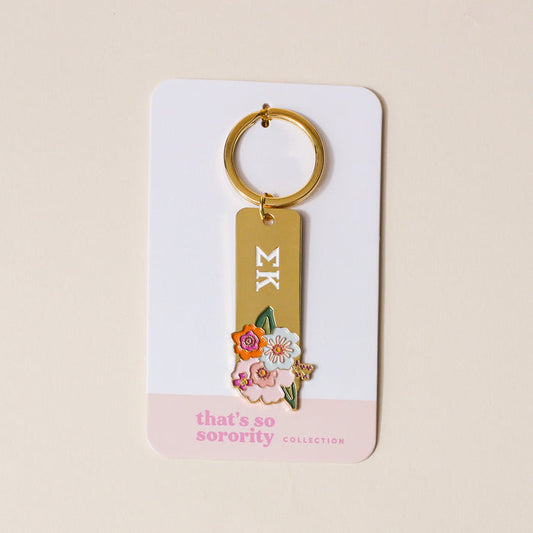 The Darling Effect’s Sigma Kappa Enamel Keychain features white ΣΚ Greek letters and colorful flowers on gold enamel, attached to a “thats so sorority COLLECTION” card—making it a perfect Sigma Kappa gift.