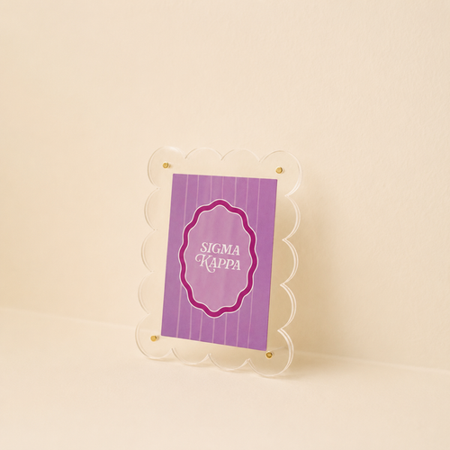 The Darling Effect’s Sigma Kappa Mini Acrylic Picture Frame Magnet features a small, scalloped-edge clear acrylic frame displaying a purple, vertically striped card with SIGMA KAPPA in white on a beige background.