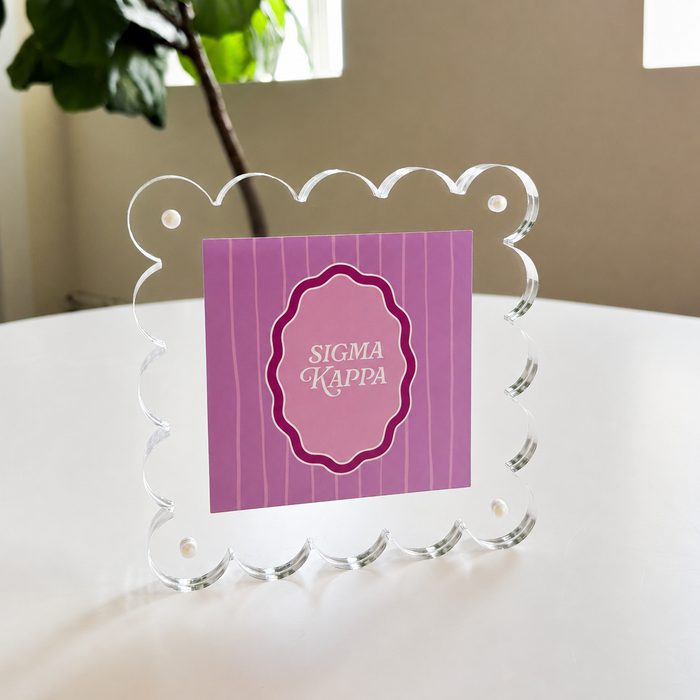 The Sigma Kappa Square Scalloped Acrylic Frame by The Darling Effect features a purple card with white lettering, displayed on a white table with greenery and natural light, adding a modern accent to your space.