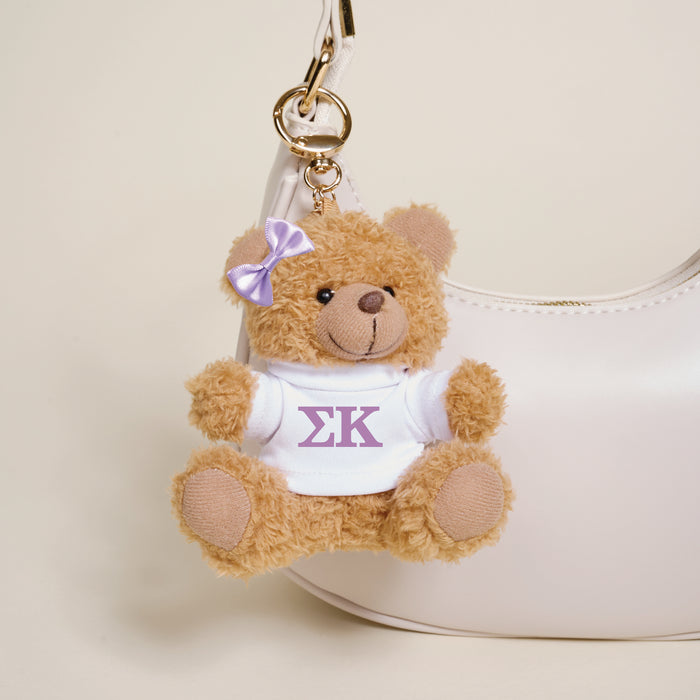 The Darling Effect's Sigma Kappa Teddy Bear Bag Charm—a small brown bear with a purple bow and white shirt featuring Greek letters—hangs from a white handbag, adding a cute sorority touch.