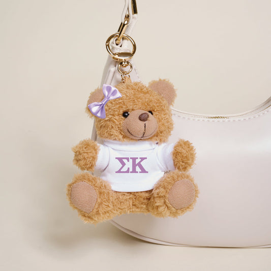 The Darling Effect's Sigma Kappa Teddy Bear Bag Charm—a small brown bear with a purple bow and white shirt featuring Greek letters—hangs from a white handbag, adding a cute sorority touch.