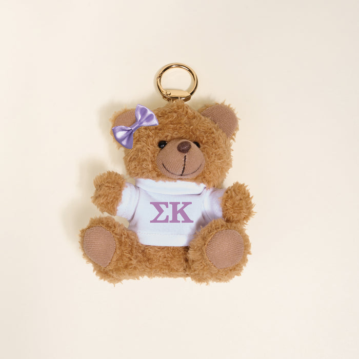 The Darling Effect Sigma Kappa Teddy Bear Bag Charm features a small bear with a lavender bow and white shirt with purple ΣΚ letters, ideal as a sorority accessory or keychain, shown on a plain light background.