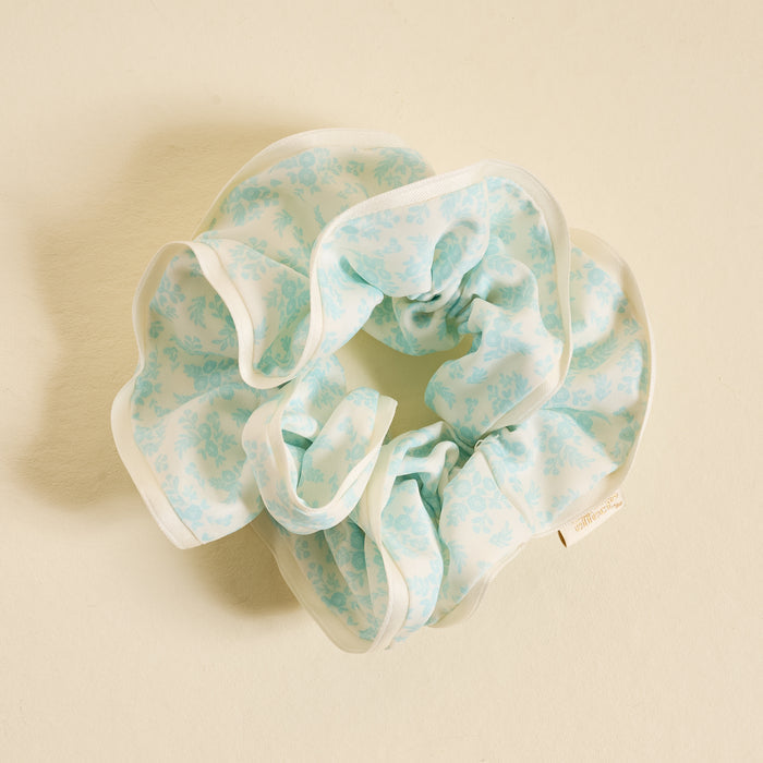 The Darling Effect's Something Blue Ruffle Up Scrunchie features light blue ruffles, floral print, and white trim—a charming hair accessory styled on a cream background to add a sweet touch to your look.
