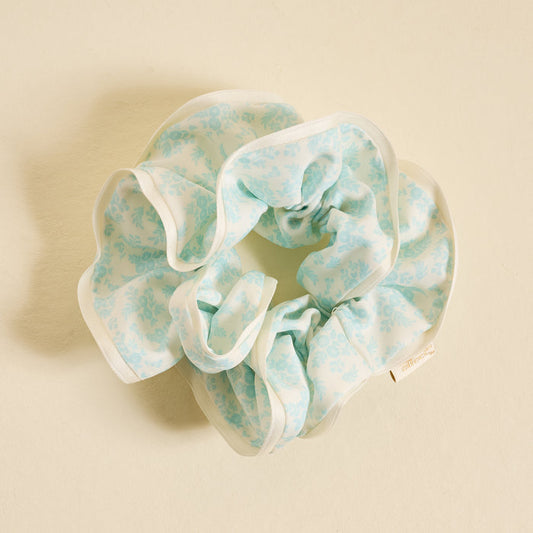 The Darling Effect's Something Blue Ruffle Up Scrunchie features light blue ruffles, floral print, and white trim—a charming hair accessory styled on a cream background to add a sweet touch to your look.