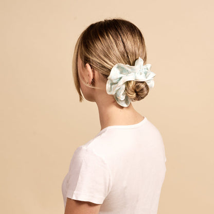 A woman with light brown hair in a low bun, secured with The Darling Effect’s Something Blue Ruffle Up Scrunchie, wears a plain white t-shirt and is shown from behind against a beige background.