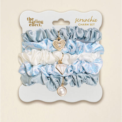 The Darling Effect’s Something Blue Scrunchie Charm Set includes light blue satin, white floral, and blue scrunchies, each with a dangling heart, diamond, or pearl charm—perfect for adding flair to any hairstyle.