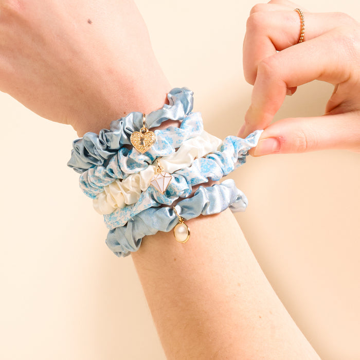 A wrist wearing The Darling Effect’s Something Blue Scrunchie Charm Set—four blue and white scrunchies, each with unique charms like a heart, geometric shape, and pearl. The other hand adjusts one of the scrunchies.
