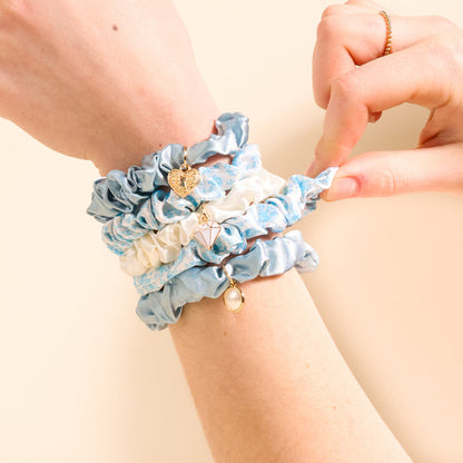A wrist wearing The Darling Effect’s Something Blue Scrunchie Charm Set—four blue and white scrunchies, each with unique charms like a heart, geometric shape, and pearl. The other hand adjusts one of the scrunchies.