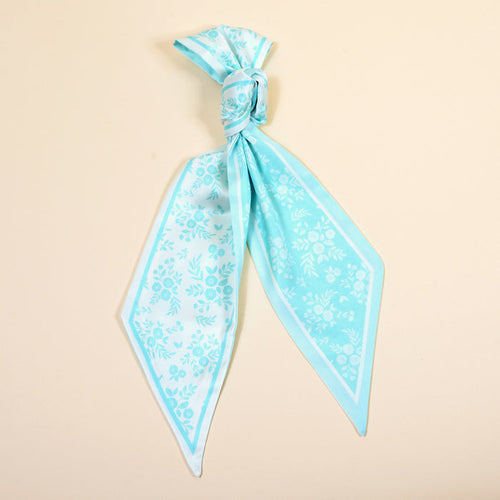 The Darling Effect’s Something Blue Twirl & Tie Scarf Accessory, featuring light blue silk with floral patterns and white borders, is styled in a loose knot against a cream background for boutique-worthy charm.