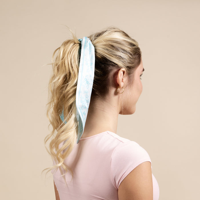 A woman with long, wavy blonde hair wears The Darling Effect’s Something Blue Twirl & Tie Scarf Accessory, tied around her ponytail. She’s seen from the back in a light pink short-sleeved shirt and gold hoops against a beige background.