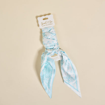The Darling Effect’s Something Blue Twirl & Tie Scarf Accessory, a light blue and white floral pet bandana, exudes boutique charm and comes with a branded tag reading "Twirl & Tie Scarf Accessory" on a beige background.