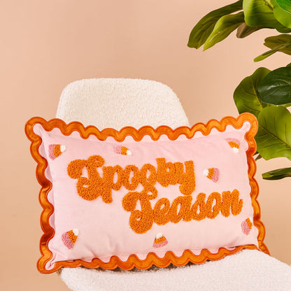 The Darling Effect’s Spooky Season Scalloped Rectangle Throw Pillow features orange text and candy corn accents, making it a playful addition to your sofa or favorite pillow set. Shown on a white chair beside a green plant.
