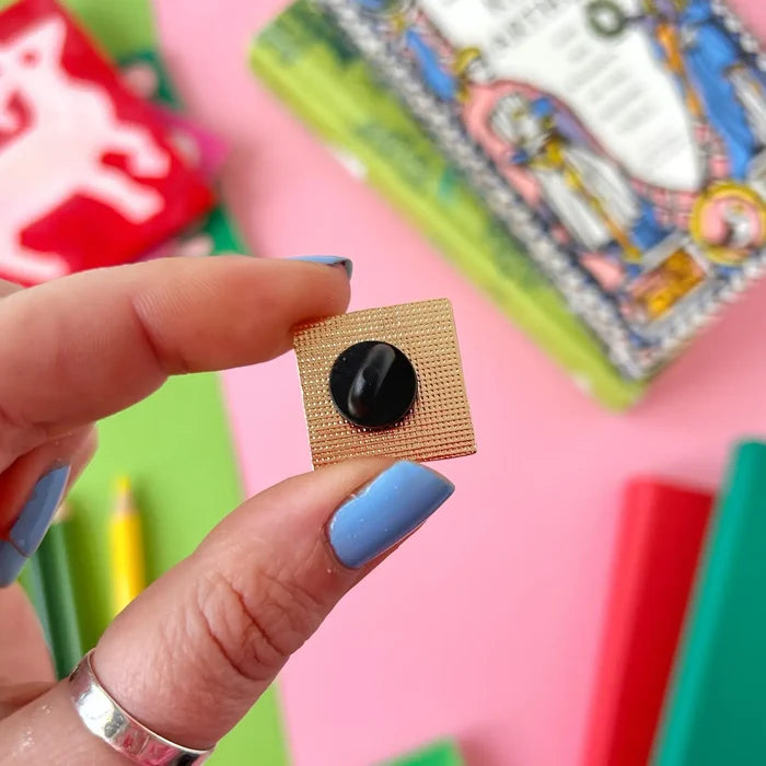 Blue-polished nails hold a Stacey McEvoy Caunt Medieval Abbey enamel pin; books and pencil visible in the stylish backdrop.