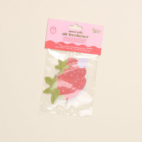 The Darling Effect’s Strawberries Sweet Ride Car Air Freshener features a strawberry shape, pink header card, and comes in a clear plastic bag, offering a refreshing scent and showing the air freshener inside.