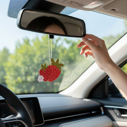 A person adjusts a rearview mirror in their car, where The Darling Effect’s Strawberries Sweet Ride Car Air Freshener hangs. Sunlight streams in, greenery glows outside, and a fresh strawberry scent lingers for the drive ahead.