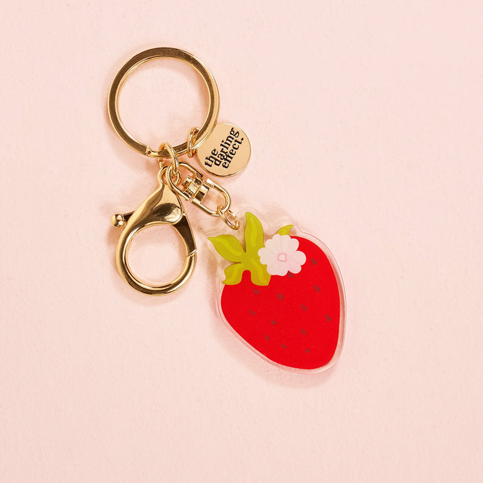 The Darling Effect's Strawberry Acrylic Keychain features a red strawberry charm, green leaves, white flower, and a round gold tag engraved with "the tiny things." This cute accessory looks lovely on a light pink background.