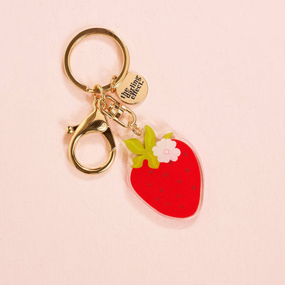 The Darling Effect's Strawberry Acrylic Keychain features a red strawberry charm, green leaves, white flower, and a round gold tag engraved with "the tiny things." This cute accessory looks lovely on a light pink background.